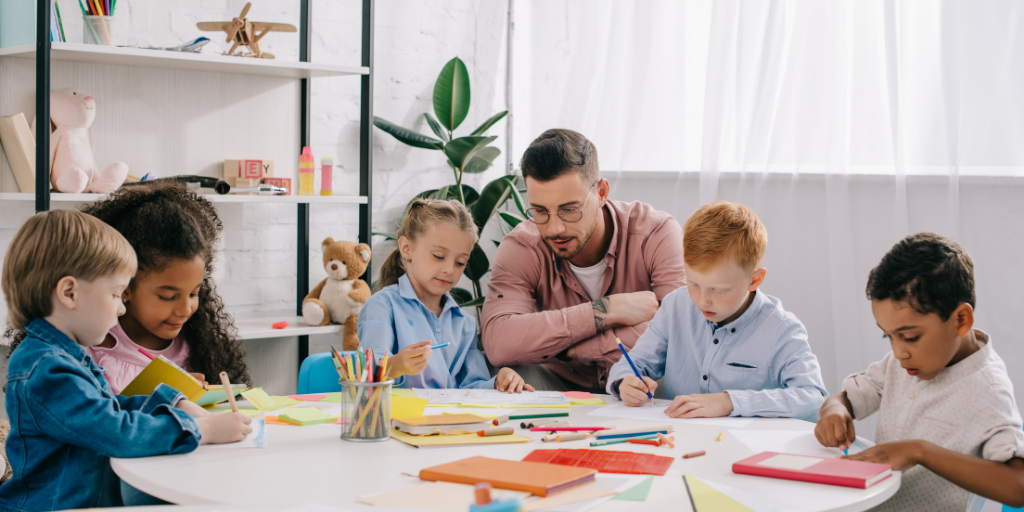 An adult sitting at a table with a group of children observing the children who are drawing and reading a book.