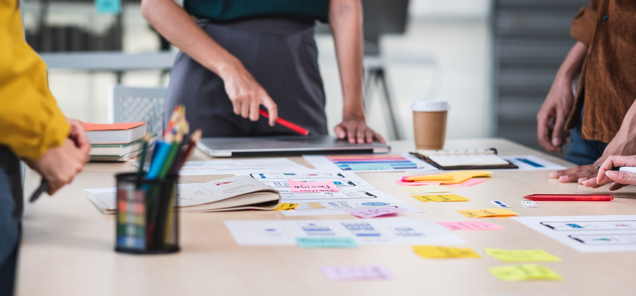 Adults standing around a desk. The desk has different colored post-it notes, a coffee cup, a lap top, books, a planner, and a cup of writing utensils on it. The adults seem to be planning. 