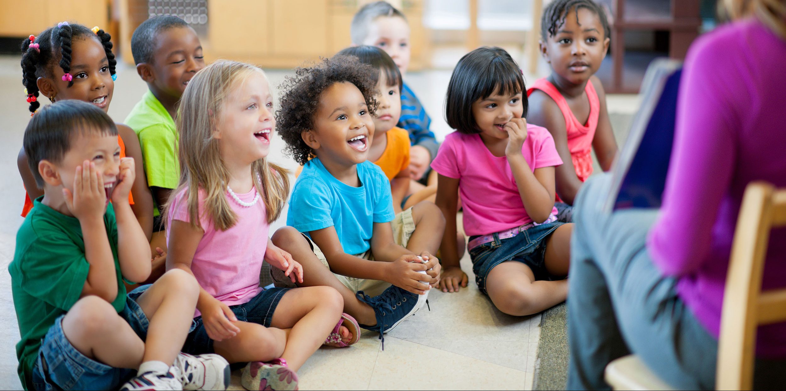 Many children sitting on the floor looking at a teacher.