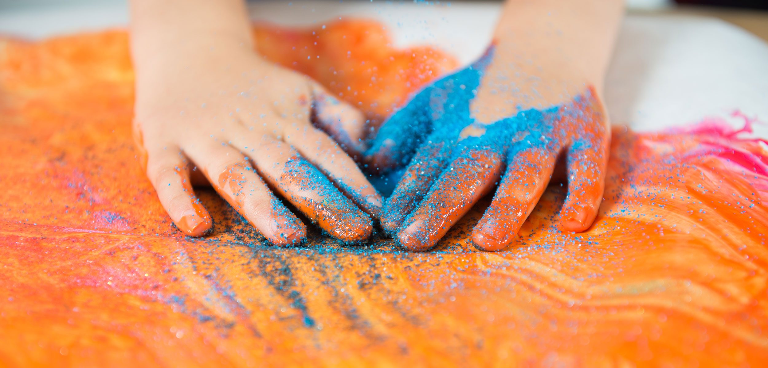 A close up of a child's hands  covered in orange paint and blue sand as they finger paint. 
