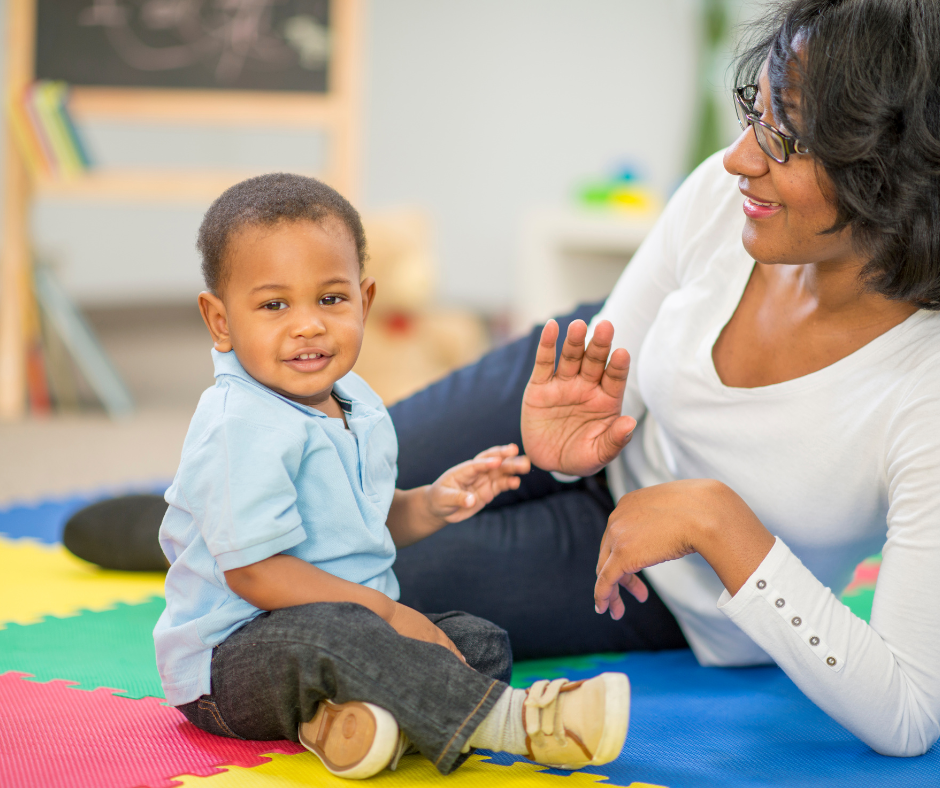 Child sitting on the floor next to an adult playing hand games.