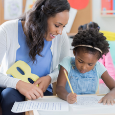 Child holding a pencil and writing and an adult observing holding a yellow uppercase letter R