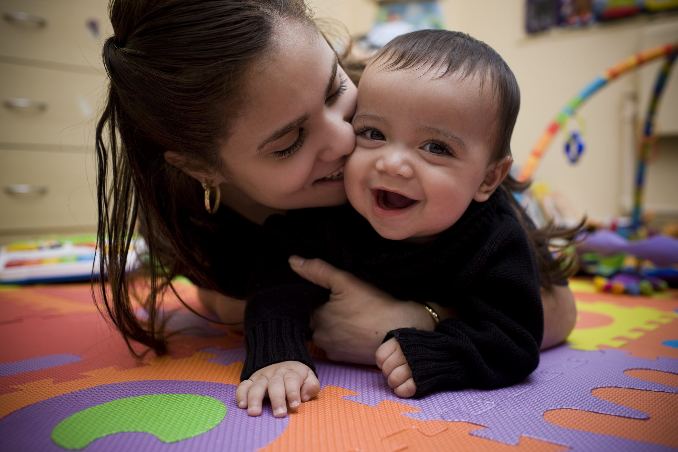 An adult hugging a smiling infant and kissing their cheek.