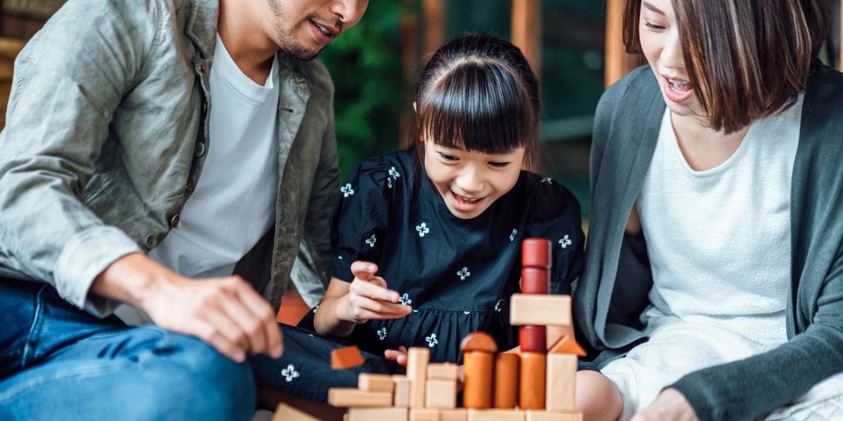 Picture of child smiling playing with stacked blocks while two adults sit on either side looking on. 