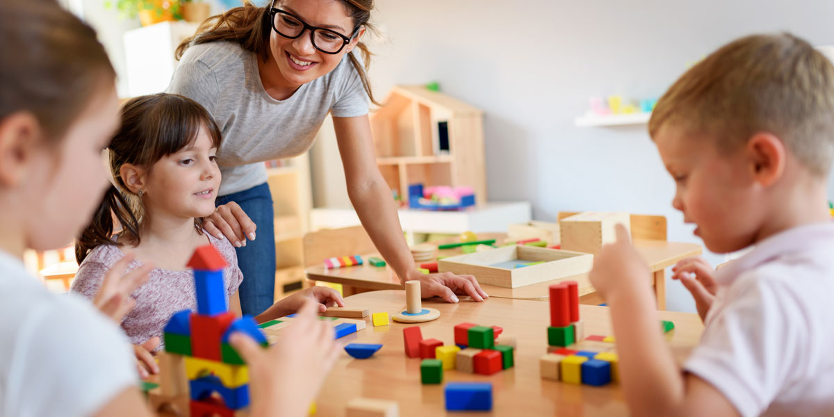 preschool teacher leaning over and smiling while three students play with blocks