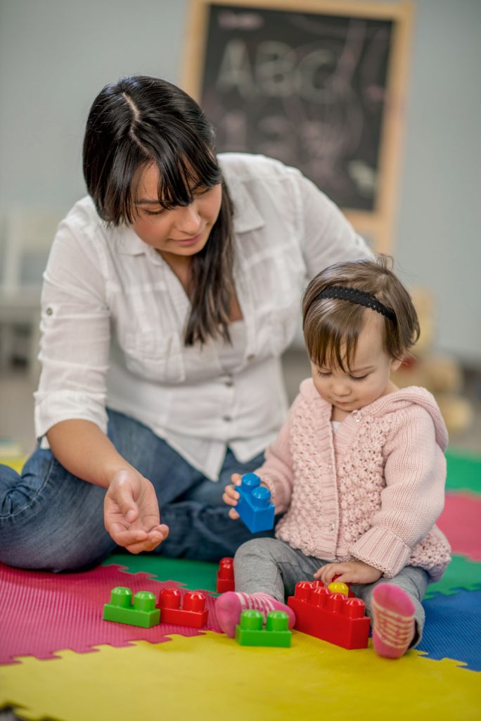An adult and a toddler sitting on the floor. The toddler is using interlocking bricks and the adult is observing.