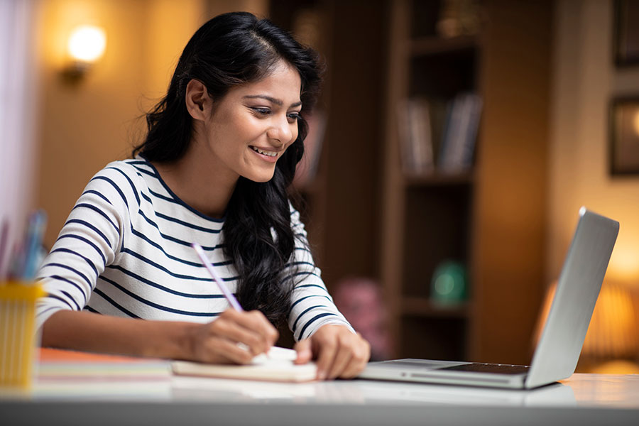 An adult looking at an open laptop screen. The adult is holding a pencil in their hand posed over paper.