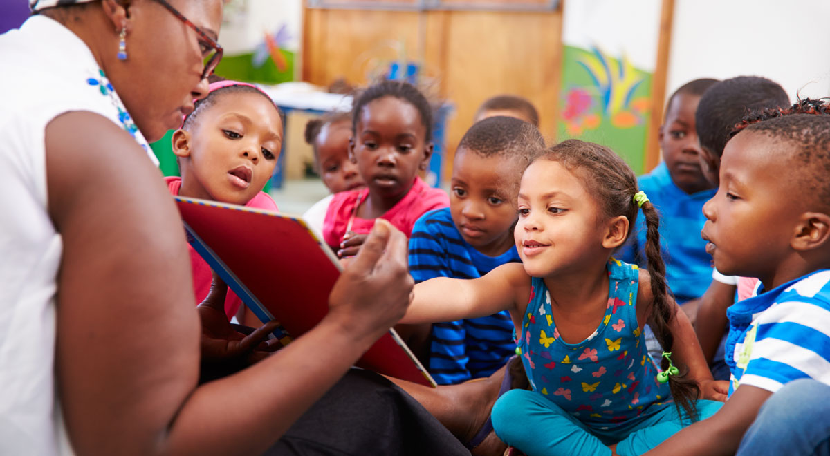 a group of African-American preschoolers sitting on the floor while a teacher reads them a book. The front girl points at the book